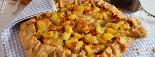 Torta salata con patate, speck e scamorza sutovaglietta a quadretti e tavolo bianco.