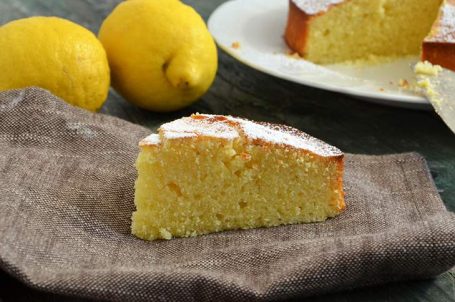 Fetta di torta al limone su canovaccio con torta intera e limoni sullo sfondo