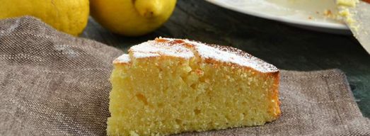 Fetta di torta al limone su canovaccio con torta intera e limoni sullo sfondo