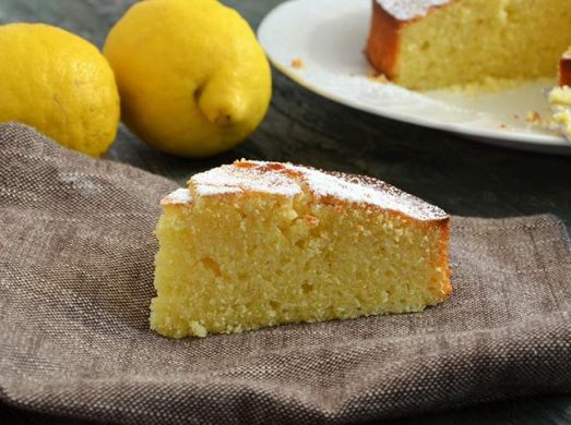 Fetta di torta al limone su canovaccio con torta intera e limoni sullo sfondo