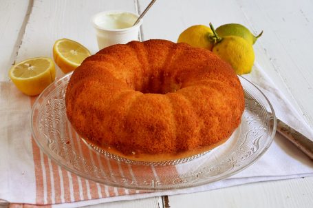 Ciambella yogurt e limone su vassoio di vetro su tovaglietta con limoni freschi tagliati