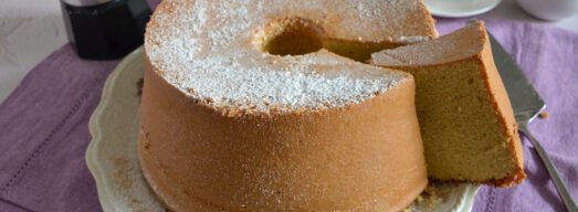 Torta su piatto da portata con fetta tagliata e dietro macchinetta del caffè e tazzine.