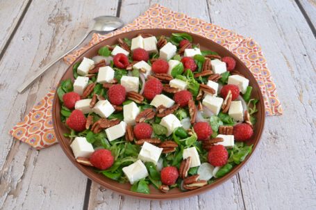 Insalata con feta a cubetti, lamponi freschi, noci pecan e valeriana servita in un piatto marrone.