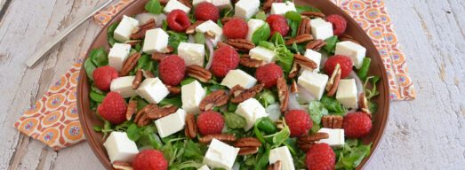 Insalata con feta a cubetti, lamponi freschi, noci pecan e valeriana servita in un piatto marrone.