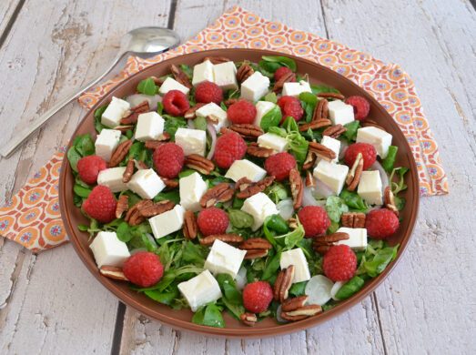 Insalata con feta a cubetti, lamponi freschi, noci pecan e valeriana servita in un piatto marrone.