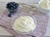 Ciotolina con crema allo yogurt su tovaglietta a fiori e ciotoline di mirtilli e yogurt sullo sfondo