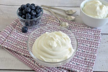 Ciotolina con crema allo yogurt su tovaglietta a fiori e ciotoline di mirtilli e yogurt sullo sfondo