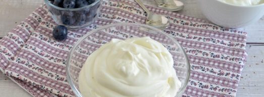 Ciotolina con crema allo yogurt su tovaglietta a fiori e ciotoline di mirtilli e yogurt sullo sfondo